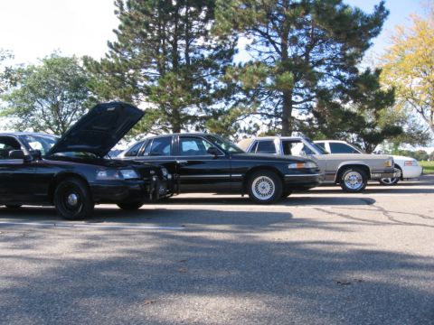 1988 Mercury Grand Marquis - Multiple vehicles, including a black police car with its hood up, are parked in a row on asphalt.