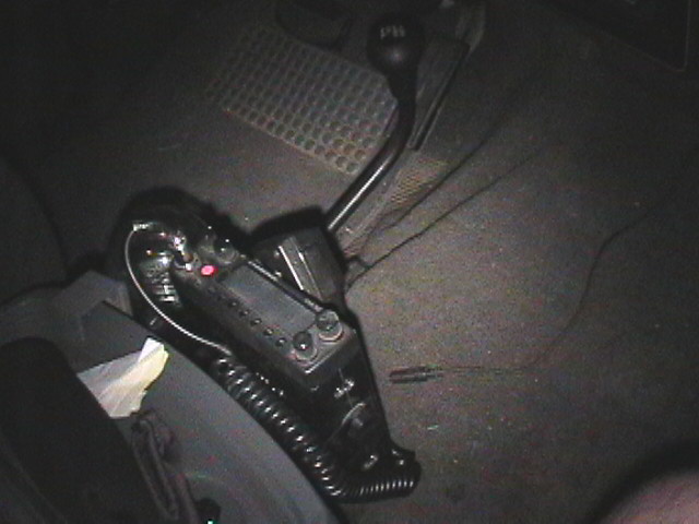 Mobile radio with coiled mic cord on carpeted floor beside a floor shifter inside a vehicle at night