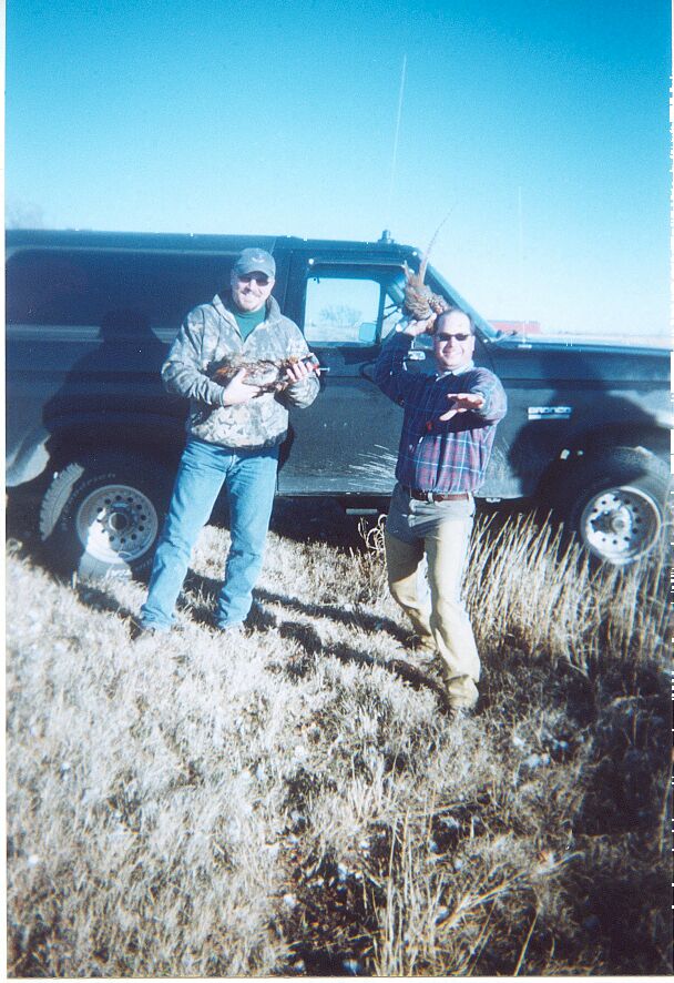 Two men in camo and flannel holding pheasants in front of a dark Ford truck in a grassy field