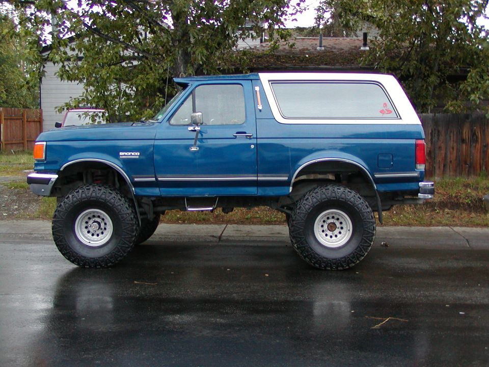 Lifted blue Ford Bronco with white top and big off-road tires, side view on wet street