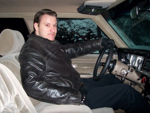 1988 Mercury Grand Marquis - Night interior shot of a young man in a black leather jacket and gloves sitting in the driver's seat of an older car with a tan/cream velou…