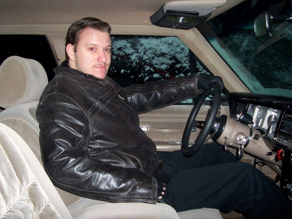 Man in black leather jacket at the wheel of a car with tan velour interior at night