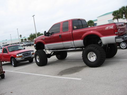 1974 Ford F-100 - A heavily modified red pickup truck with oversized tires is parked in a lot next to a smaller red truck.