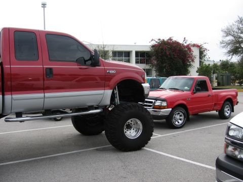 1974 Ford F-100 - A lifted red pickup truck with massive tires is parked next to a red Ford Ranger, with a building in the background.