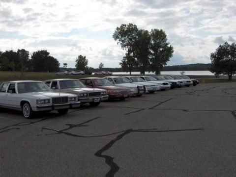 1988 Mercury Grand Marquis - Long row of full-size sedans, mostly Ford and Mercury, parked at a paved lakeside lot under a cloudy sky at a club meet.