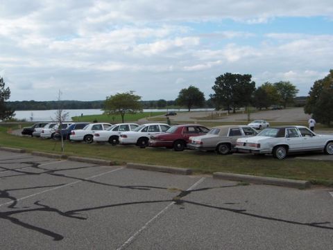 1988 Mercury Grand Marquis - Several sedans are parked in a row next to a body of water on an overcast day.