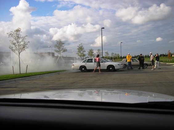 White sedan doing a burnout in a parking lot with people watching