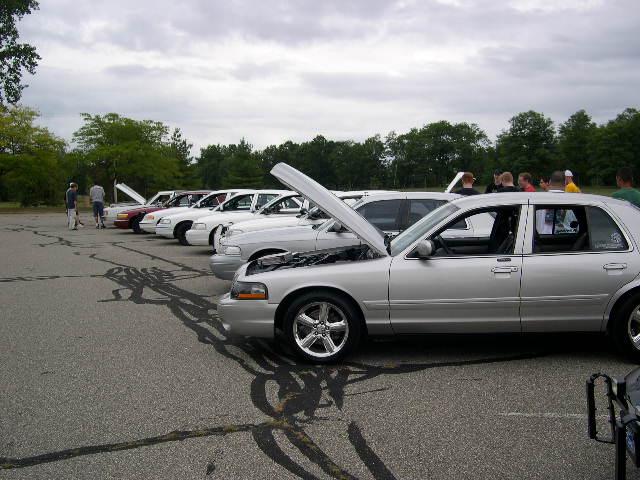 Several silver sedans parked in a lot, some with hoods open. People are standing around them