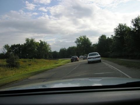 1988 Mercury Grand Marquis - A white 1988 Mercury Grand Marquis leads a line of cars on a two-lane road under a cloudy sky.