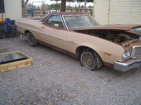 1974 Ford Ranchero - A tan 1974 Ford Ranchero sits with a flat front tire and its hood open, parked on a gravel lot.