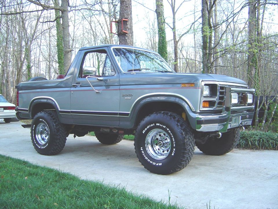 Two-tone gray Ford Bronco, top off, lifted on chrome wheels in a wooded driveway