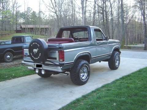 1986 Ford Bronco - Three-quarter rear view of a gray two-tone full-size Ford Bronco with the top removed and a red interior, parked on a concrete driveway in…