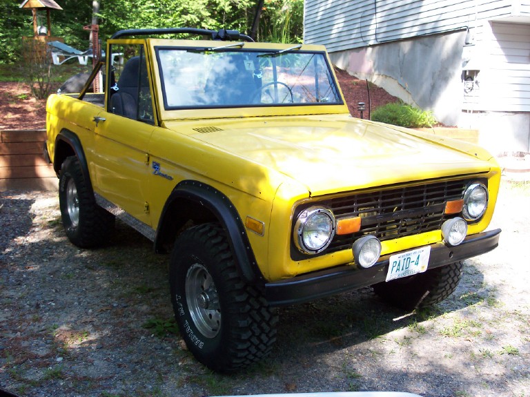 Yellow topless early Ford Bronco with roll bar, fender flares, and off-road tires, front plate reading PAID-4
