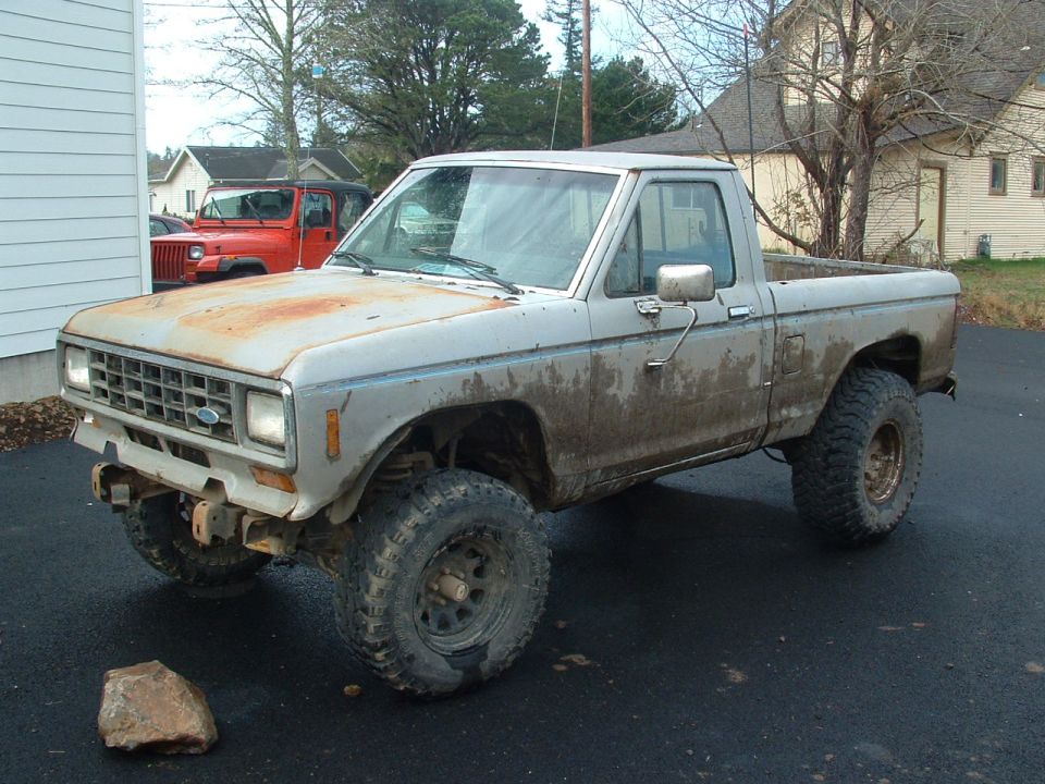 Lifted silver 1983 Ford Ranger pickup with rusty hood and big mud tires, parked on asphalt driveway