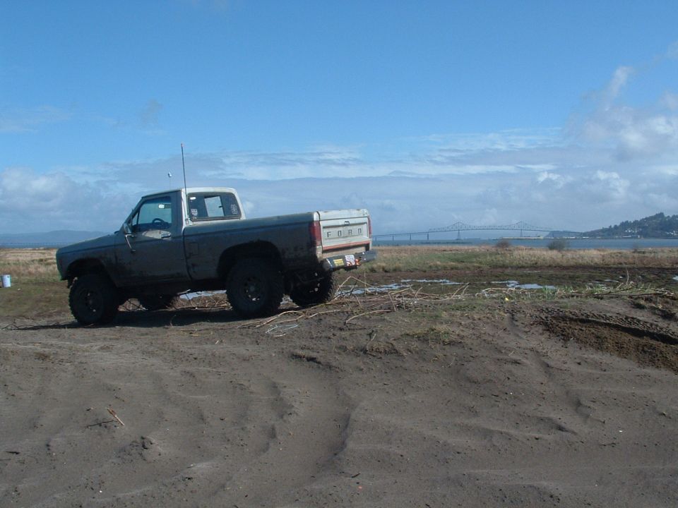 Lifted primer-gray 1980s Ford pickup parked on a sandy flat with a bridge behind