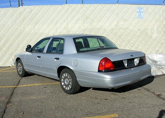 Silver Ford Crown Victoria sedan, rear three-quarter view, parked by a corrugated fence with snow nearby