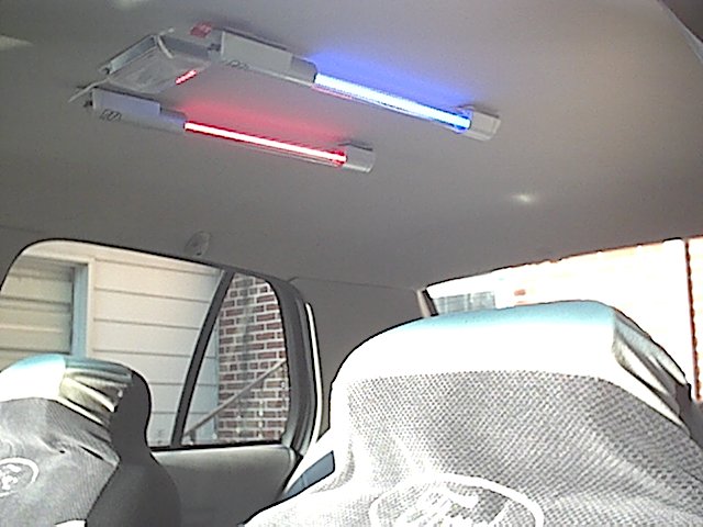 Car interior ceiling with red and blue LED tube lights mounted to gray headliner above seat-covered front seats
