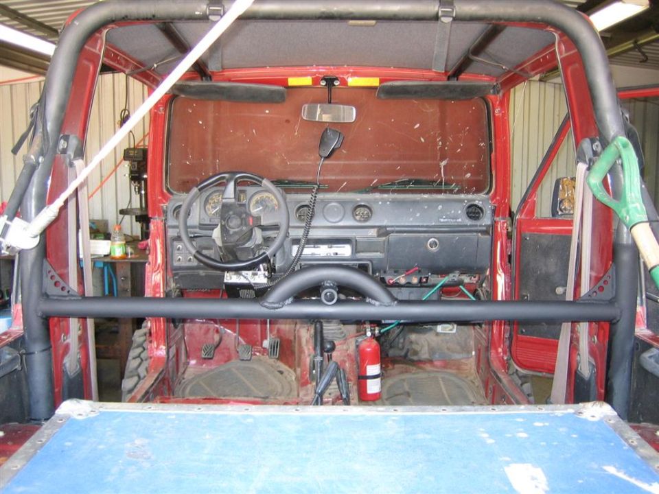 Interior of a red 1987 Suzuki Samurai showing dashboard, steering wheel, roll cage, and fire extinguisher
