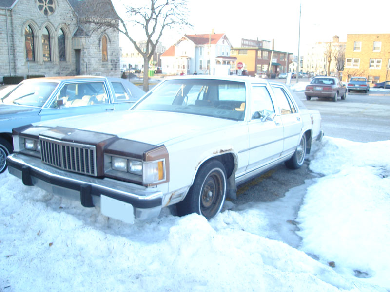 White sedan with brown hood in snow. Other cars and buildings in background
