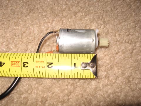 1990 Ford Bronco - A small bare-metal brushed DC motor with black wire leads, orange capacitors and a white nylon pinion gear, laid on beige carpet next to a…