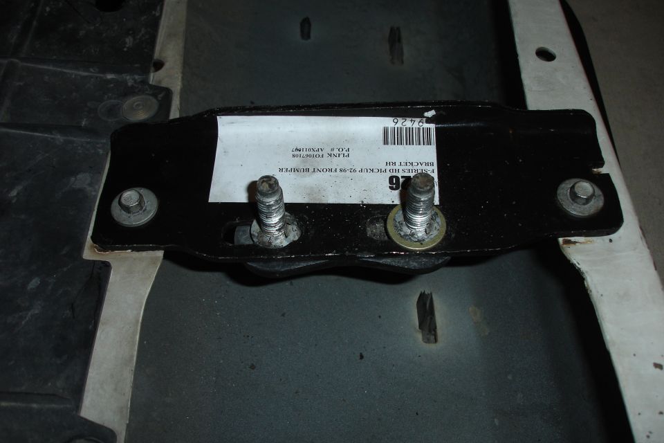 Close-up of a black metal bracket with two bolts, a white label, and a barcode. Label reads 'E-SERIES HD PICKUP 92-96 FRONT BUMPER BRACKET KIT'