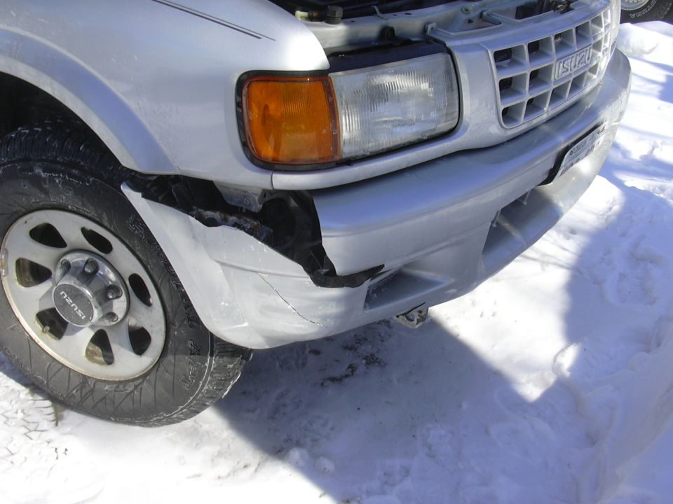 Front corner of a silver Isuzu Rodeo with a damaged bumper and fender. Snow on the ground