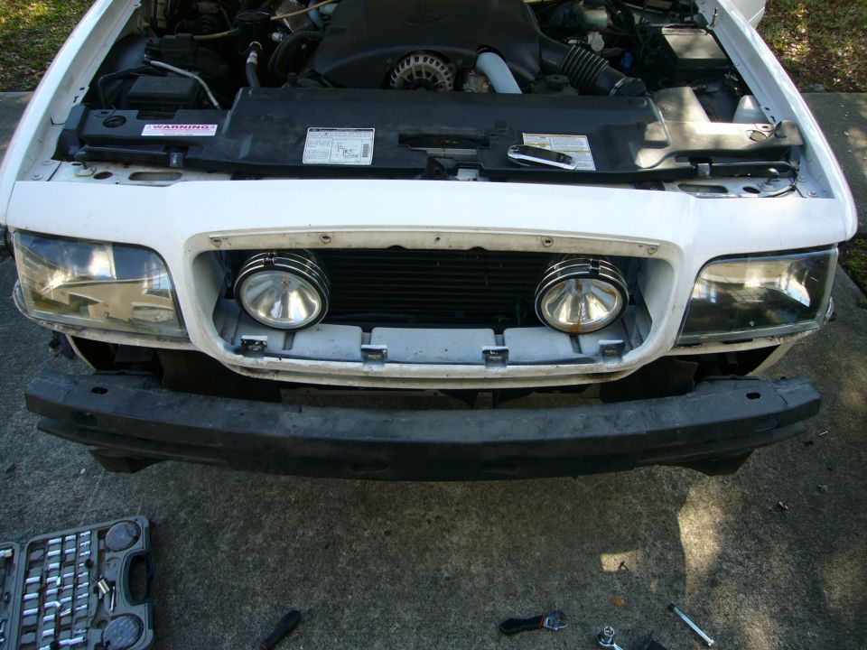 Front of a 2000 Ford Crown Victoria with hood open, showing engine and grille with auxiliary lights. Bumper removed, tools nearby