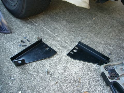 2000 Ford Crown Victoria - Two black metal brackets, likely for a Go Rhino push bumper, lie on a concrete surface next to a tire and a tool case.