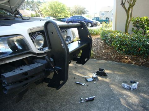2000 Ford Crown Victoria - A push bumper is being installed on the front of a white vehicle, with various parts and tools scattered on the pavement.