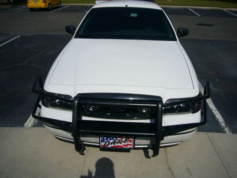 2000 Ford Crown Victoria - Elevated head-on view of a white Ford Crown Victoria parked in a sunlit lot with a fully installed black push bar and wing-style headlight…