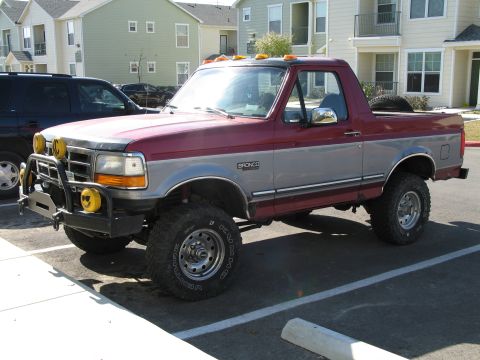 1995 Ford Bronco - Front three-quarter view of a lifted two-tone red-over-gray Ford Bronco with its hardtop removed, parked in an apartment lot. It wears a tu…