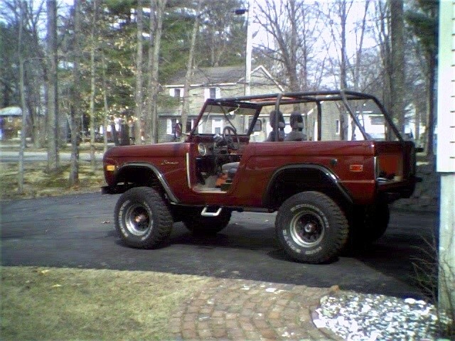 Maroon early Ford Bronco, side view, doorless with hardtop, lifted on big tires in driveway