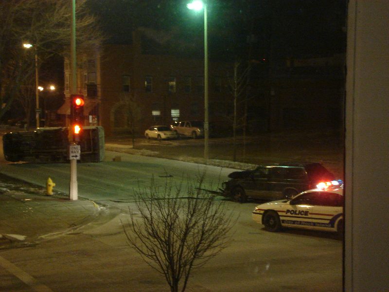 Overturned car and police cars at night, with traffic lights and buildings in background