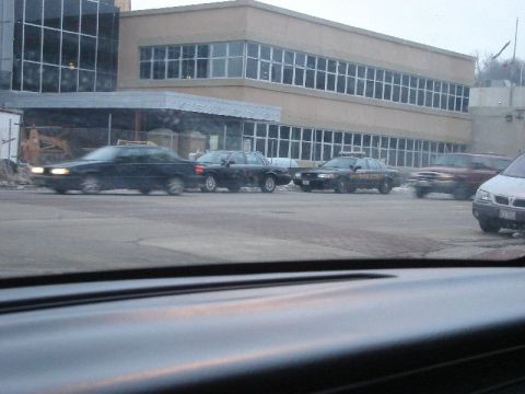 1992 Ford Crown Victoria - A line of vehicles, including a police car, are parked in front of a modern building.