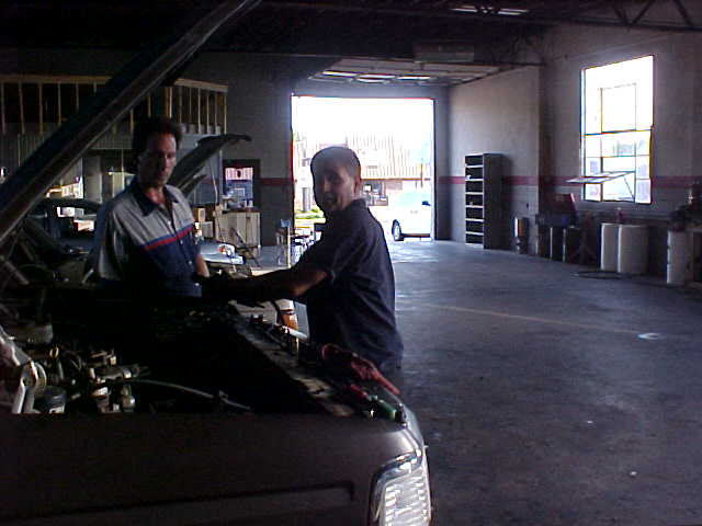 Two men working under the raised hood of a truck inside a dim garage bay