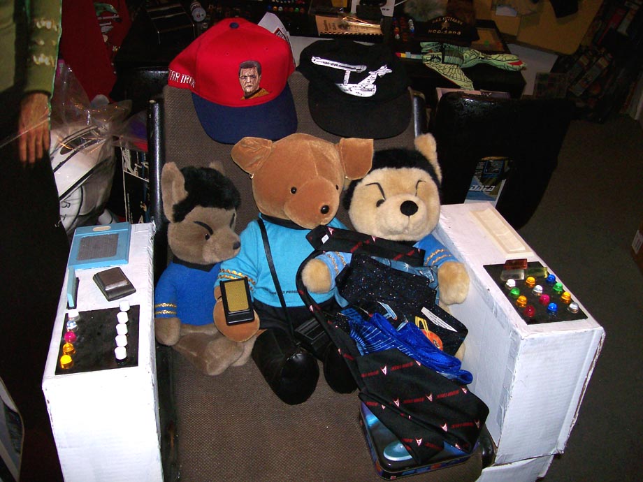 Three stuffed animals dressed in Star Trek uniforms sit on a chair with Star Trek hats and memorabilia
