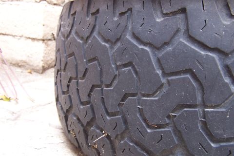 1990 Ford Bronco - A close-up of a tire tread shows a worn chevron pattern against a blurred background of light-colored bricks and some foliage.