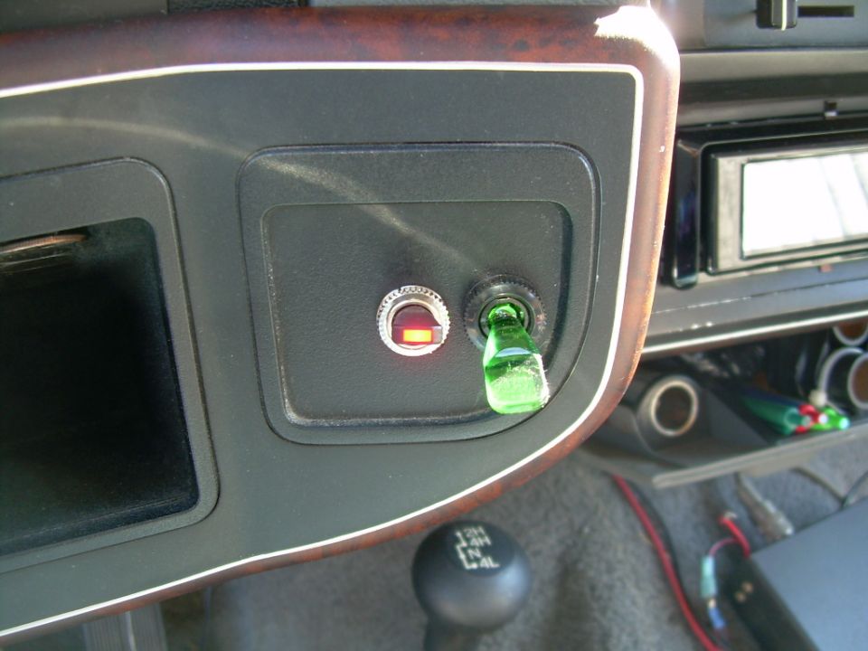 Dashboard interior showing two switches, one with a red light, and a green bottle cap on the other. Gear shifter is visible below