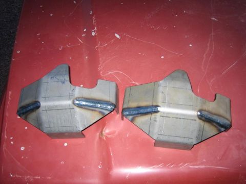 1987 Suzuki Samurai - Two custom-fabricated metal brackets are shown on a red surface, indicating they are parts for a vehicle modification.