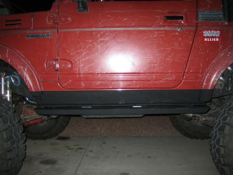 1987 Suzuki Samurai - Side view of a red off-road vehicle with prominent sliders and body damage. A "ROCKATHON ALLIED" sticker is present on the door.