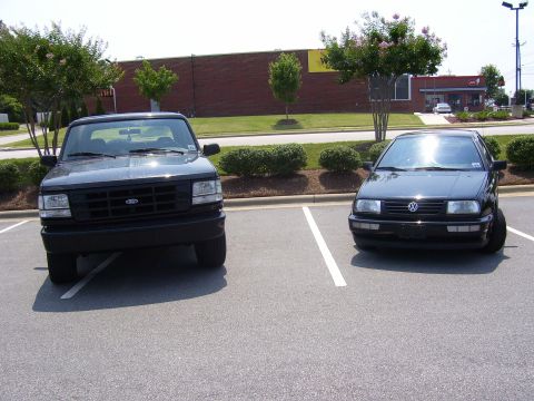 1996 Ford Bronco - Two dark-colored vehicles are parked side-by-side in an outdoor parking lot.