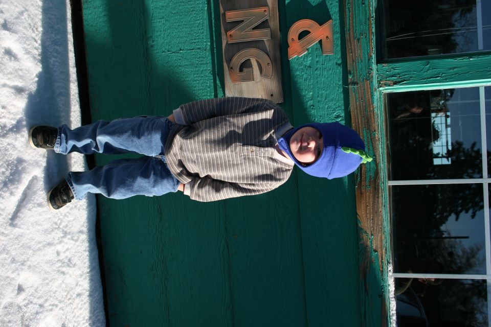 Kid in blue hat and jeans standing in snow next to a green building