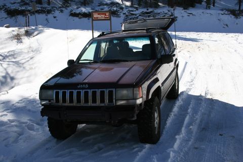 1996 Ford Bronco - A maroon Jeep Grand Cherokee is parked on a snow-covered road, with its rear hatch open, near a sign indicating a recreation area.