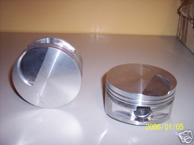 1972 Ford Ranchero - Two pistons are displayed on a flat surface, with one angled to reveal a diagonal cut on its crown.