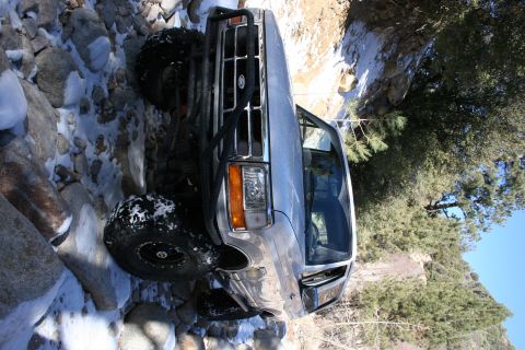1996 Ford Bronco - A modified Ford Bronco navigates a rocky, snow-dusted trail with trees and a clear blue sky in the background.