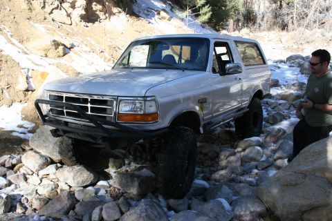 1996 Ford Bronco - A man stands by a silver 1996 Ford Bronco as it navigates a rocky, off-road trail.