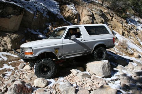 1996 Ford Bronco - A white and silver 1996 Ford Bronco tackles a challenging off-road trail with snow and rocks.