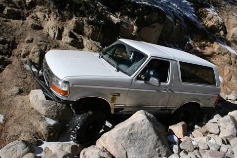 1996 Ford Bronco - A white 1996 Ford Bronco tackles a challenging off-road course, with its tires positioned on large rocks.