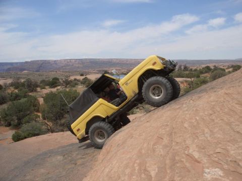 1975 Ford Bronco - A yellow off-road vehicle navigates a challenging rocky incline under a clear blue sky.