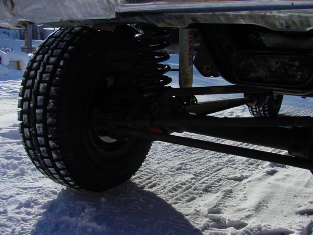 Underside of lifted truck on snow showing studded tire, coil spring and front axle